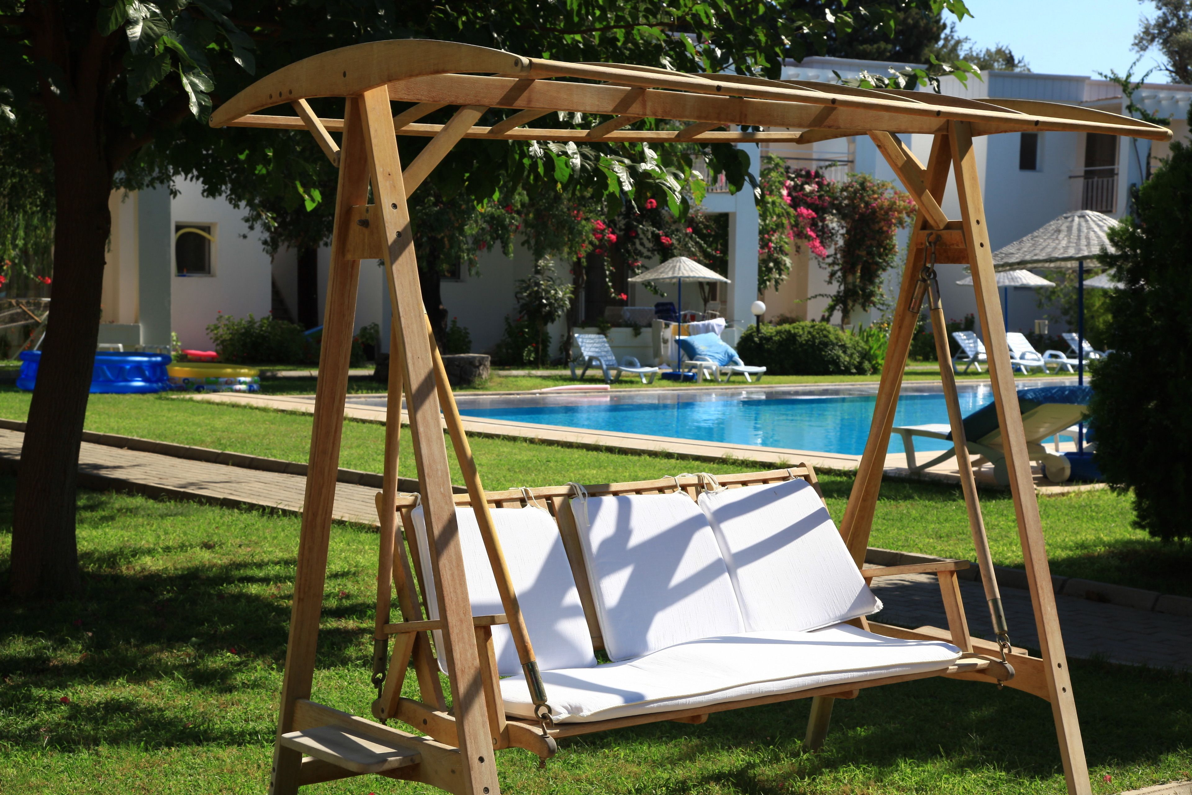 A wooden swing chair with white cushions by a swimming pool, surrounded by a well-maintained lawn and lounge chairs.