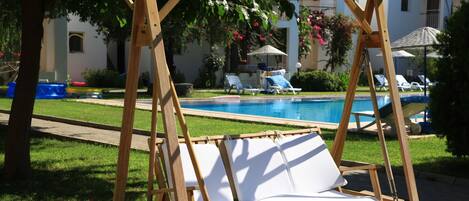A wooden swing chair with white cushions by a swimming pool, surrounded by a well-maintained lawn and lounge chairs.