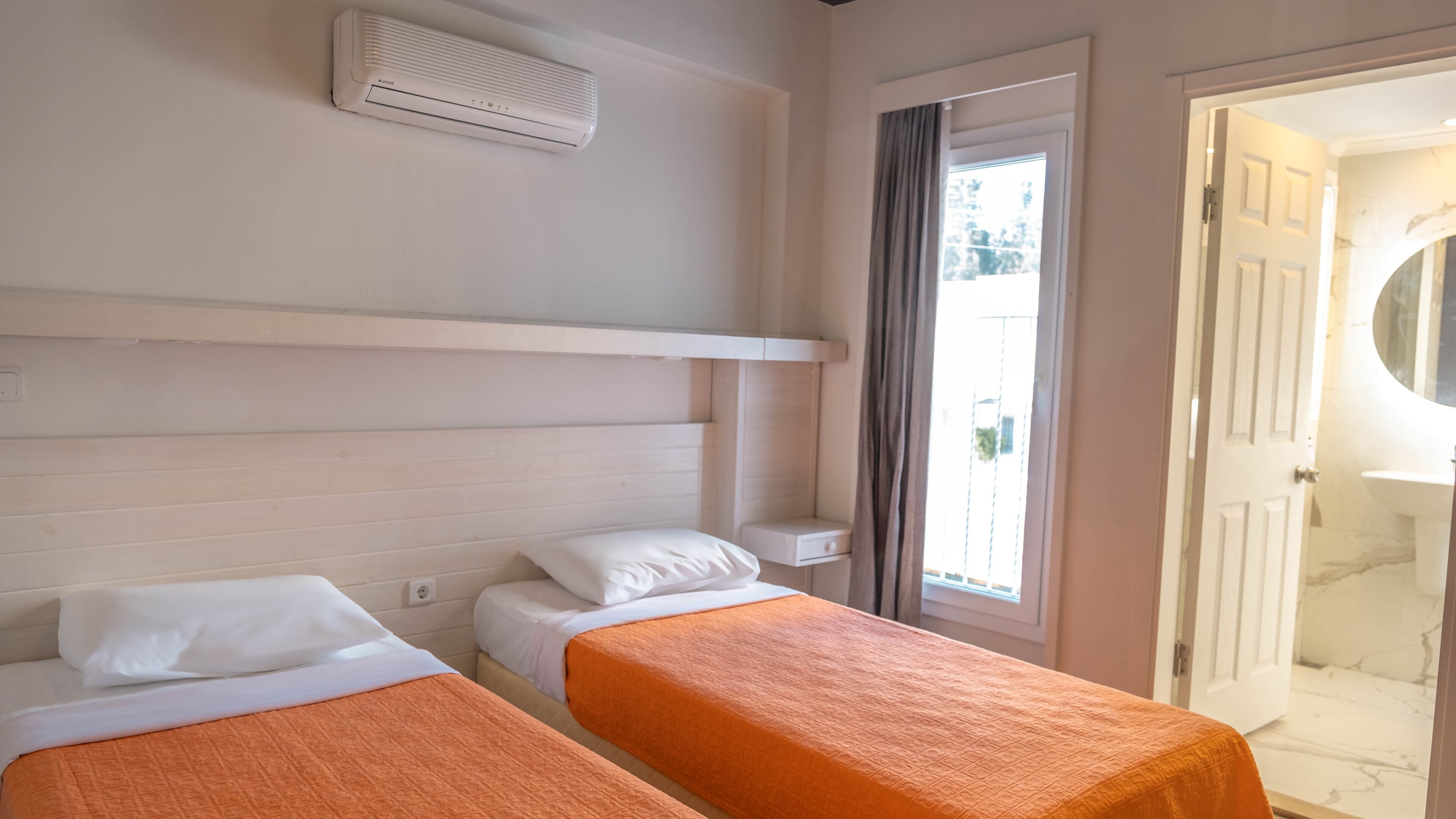 A hotel room with two single beds, a window with curtains, a bathroom with a mirror, and a white air conditioning unit on the wall.