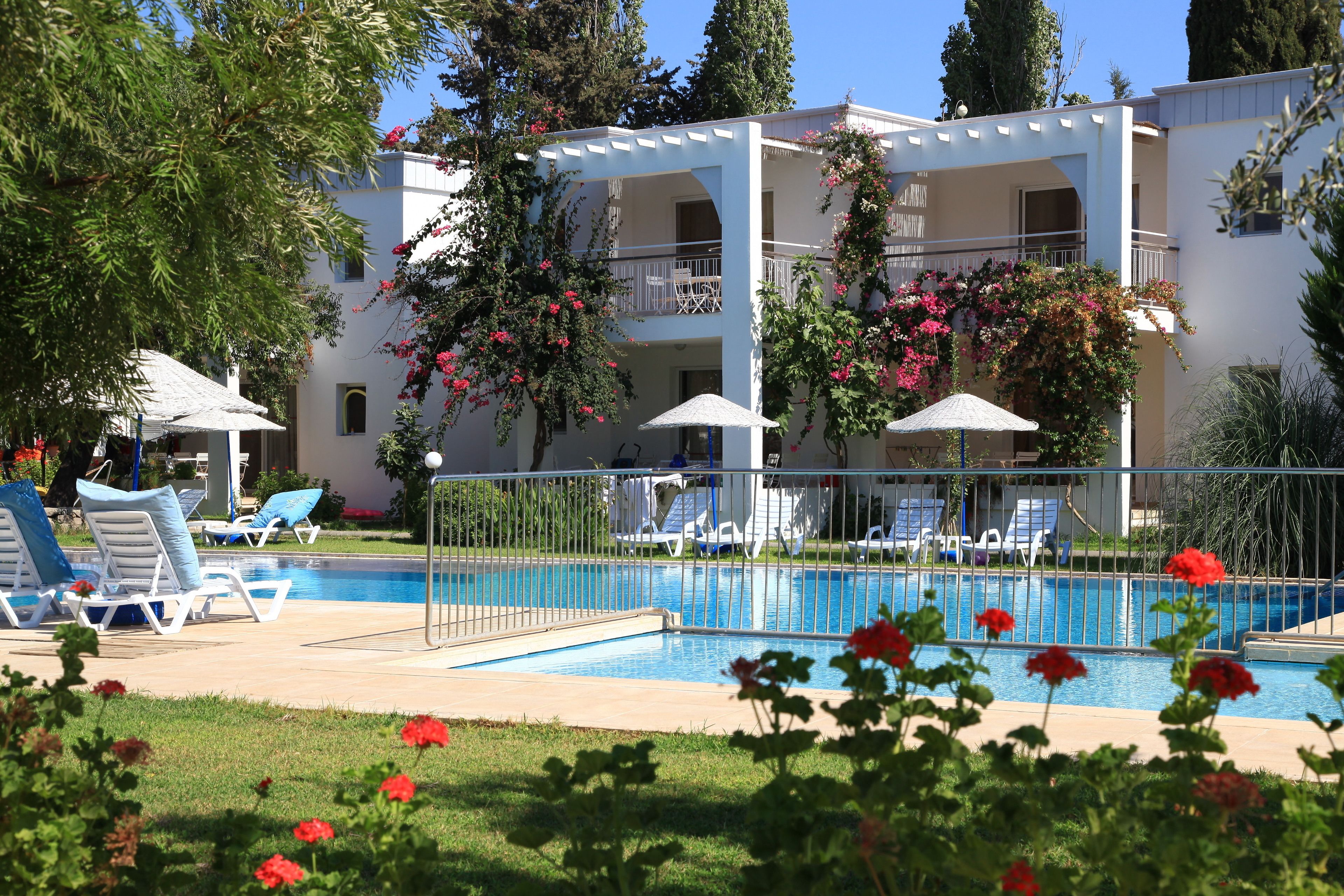 Zona de piscina con sillas de descanso y sombrillas, rodeada por un edificio blanco con balcones y plantas en flor.