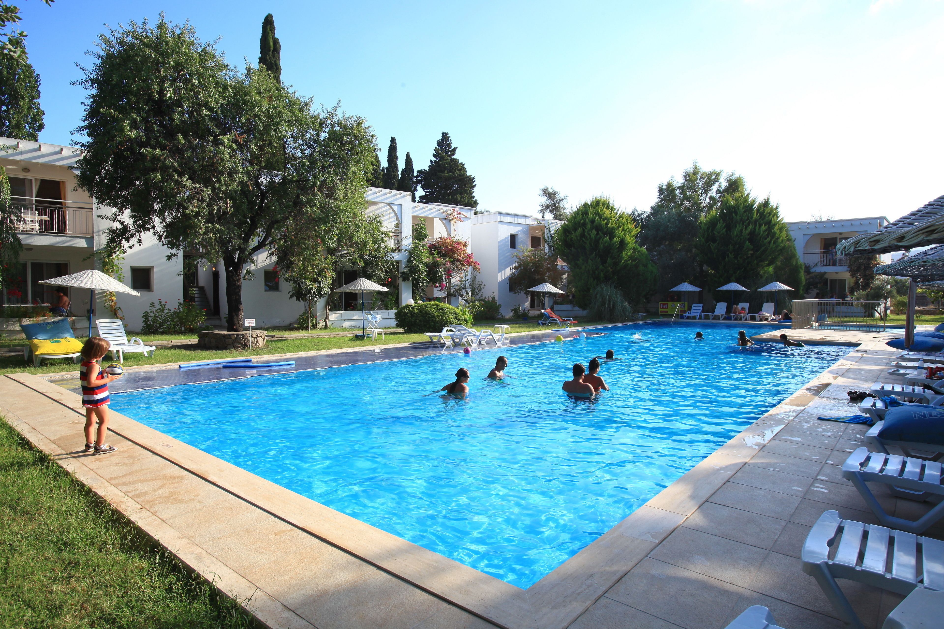 A swimming pool surrounded by lounge chairs and umbrellas, with people swimming and others relaxing.