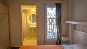 Una habitación de hotel con una cama, un ventanal con vistas a árboles, un baño con lavamanos y espejo, y una puerta que da a otra habitación.