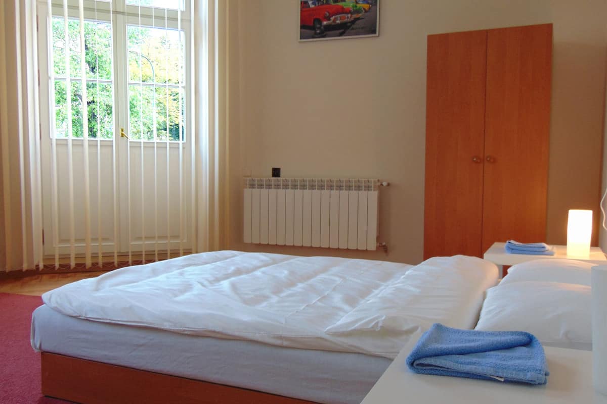 A bedroom with a bed, a radiator, a window with blinds, a wooden wardrobe, and a small table with a lamp and towels.