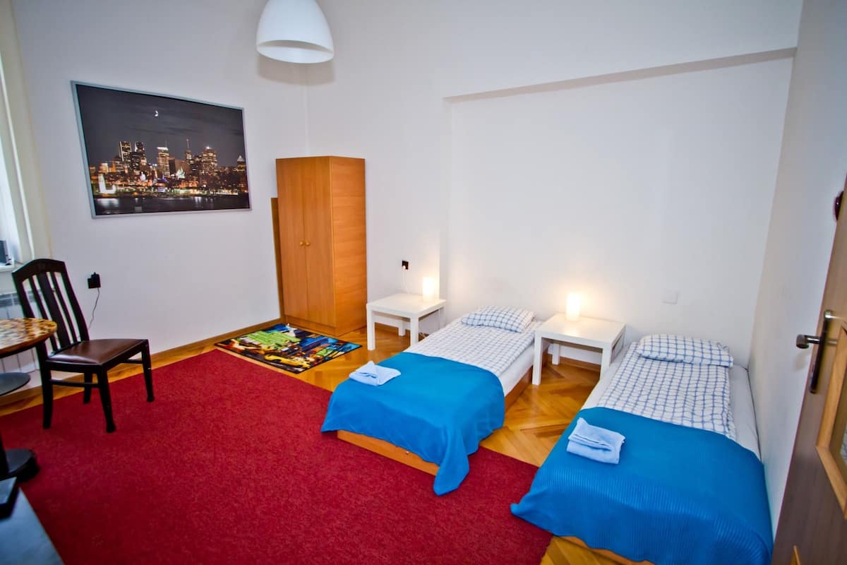 A room with two beds, a red carpet, a wooden wardrobe, a chair, and a cityscape painting on the wall.
