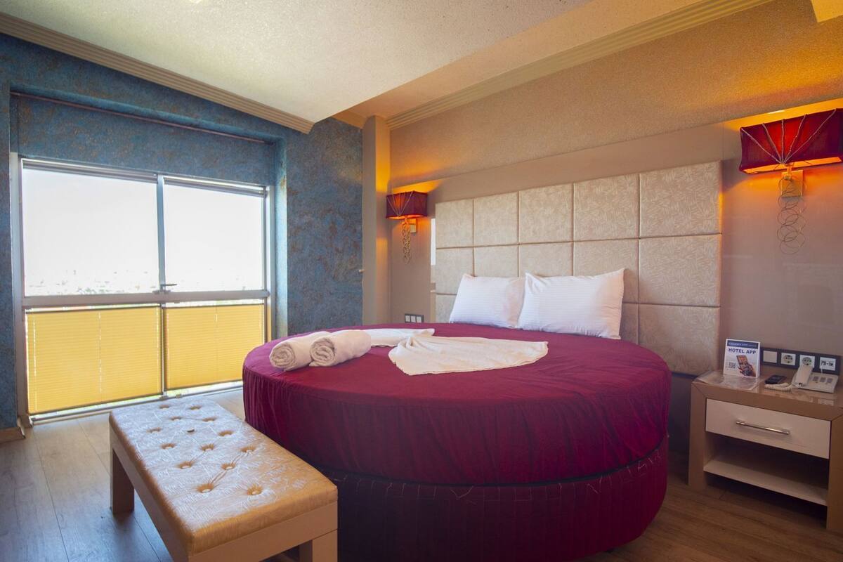A hotel room with a large bed, a bedside table, a bench, a window, and a wall-mounted phone.