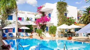 A swimming pool with lounge chairs and umbrellas, surrounded by white buildings with blue shutters and balconies. - Galini Apartments (Hersonissos)