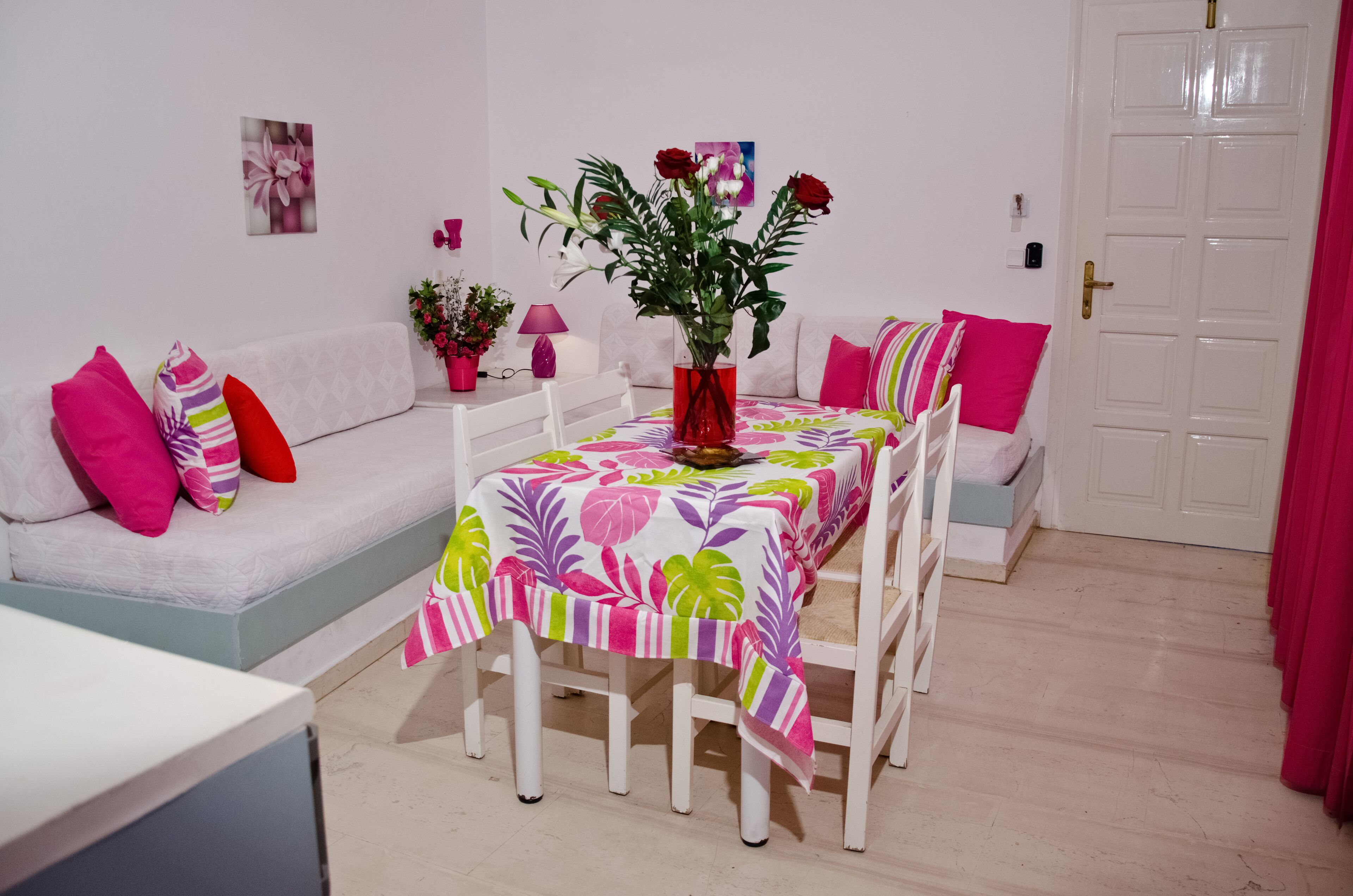 Un coin repas avec une table dressée pour quatre personnes, entourée de chaises blanches. Un vase rouge contenant des fleurs est posé sur la table.