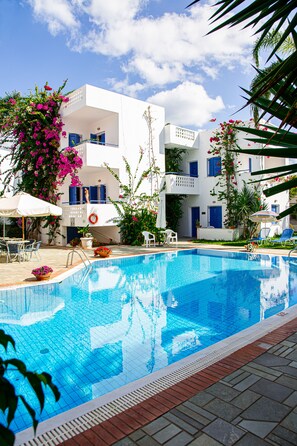 A swimming pool with a white building in the background, surrounded by plants and outdoor furniture. - Galini Apartments (Hersonissos)