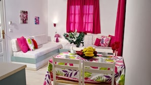 A cozy living room with a sofa, a dining table with chairs, and a window with pink curtains. - Galini Apartments (Hersonissos)