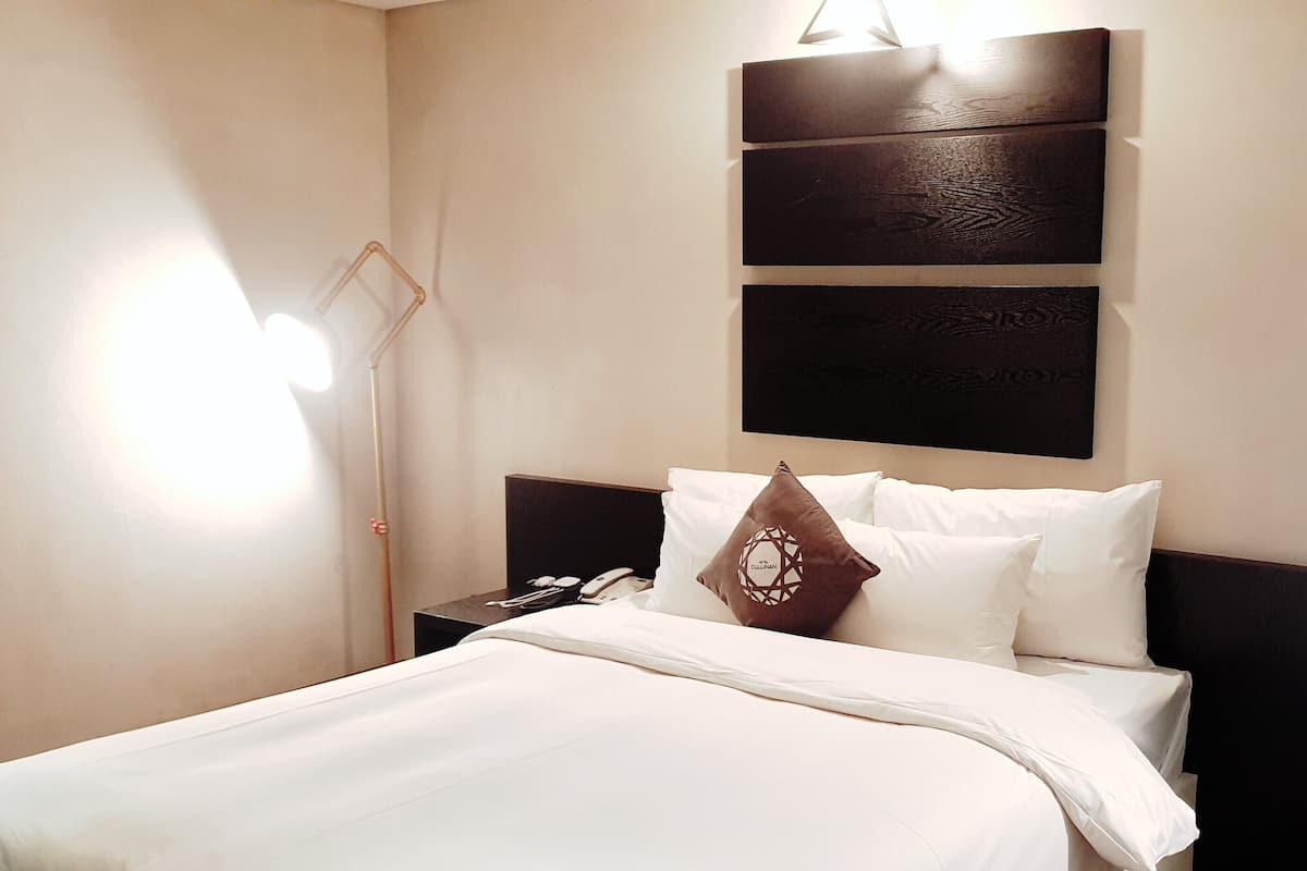 Una cama bien hecha con sábanas blancas, un cojín decorativo marrón y un cabecero con tres paneles oscuros. Hay dos lámparas de noche con pantallas redondas y una lámpara de pared.