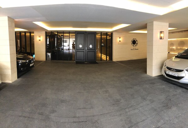 A hotel entrance with a black reception desk, a white car parked on the right, and a black car on the left.