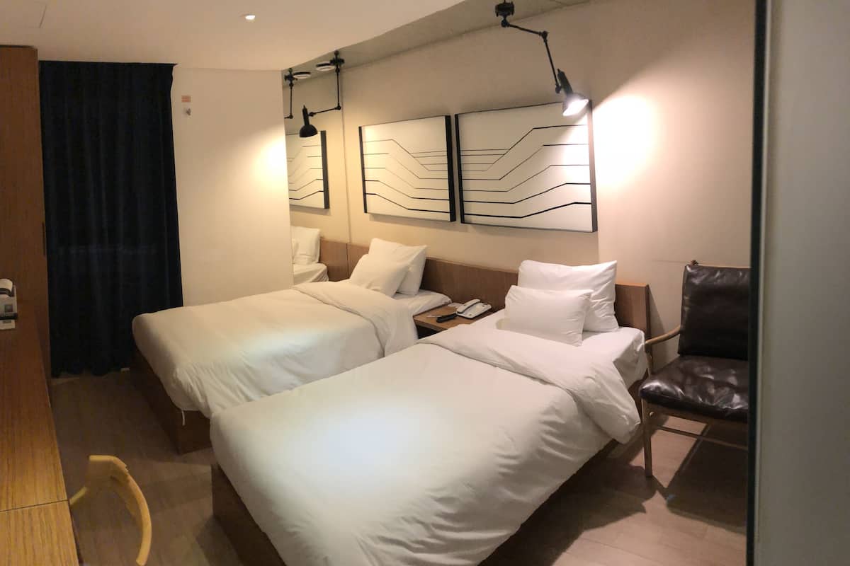 Habitación de hotel con dos camas individuales, un escritorio de madera, una silla y una lámpara fijada a la pared.