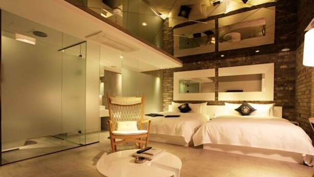 Una habitación de hotel moderna con pared de vidrio, cama, sillón y una pequeña mesa.