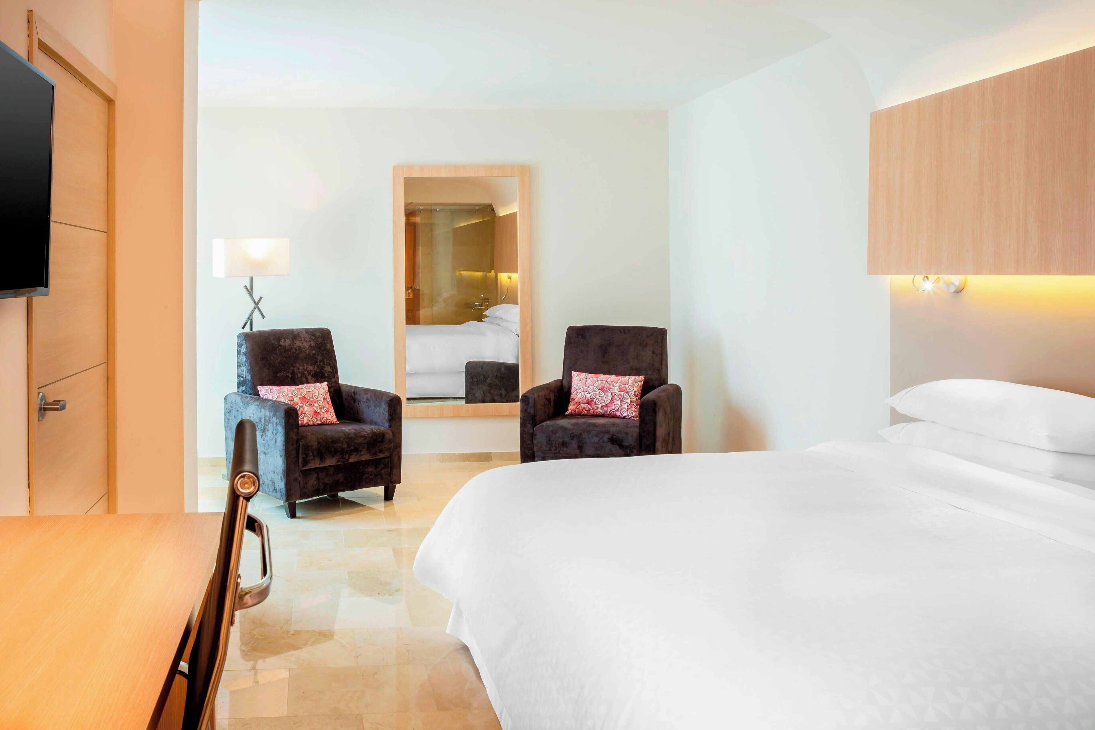 Une chambre d’hôtel avec un grand lit, deux fauteuils, un téléviseur à écran plat et un miroir.