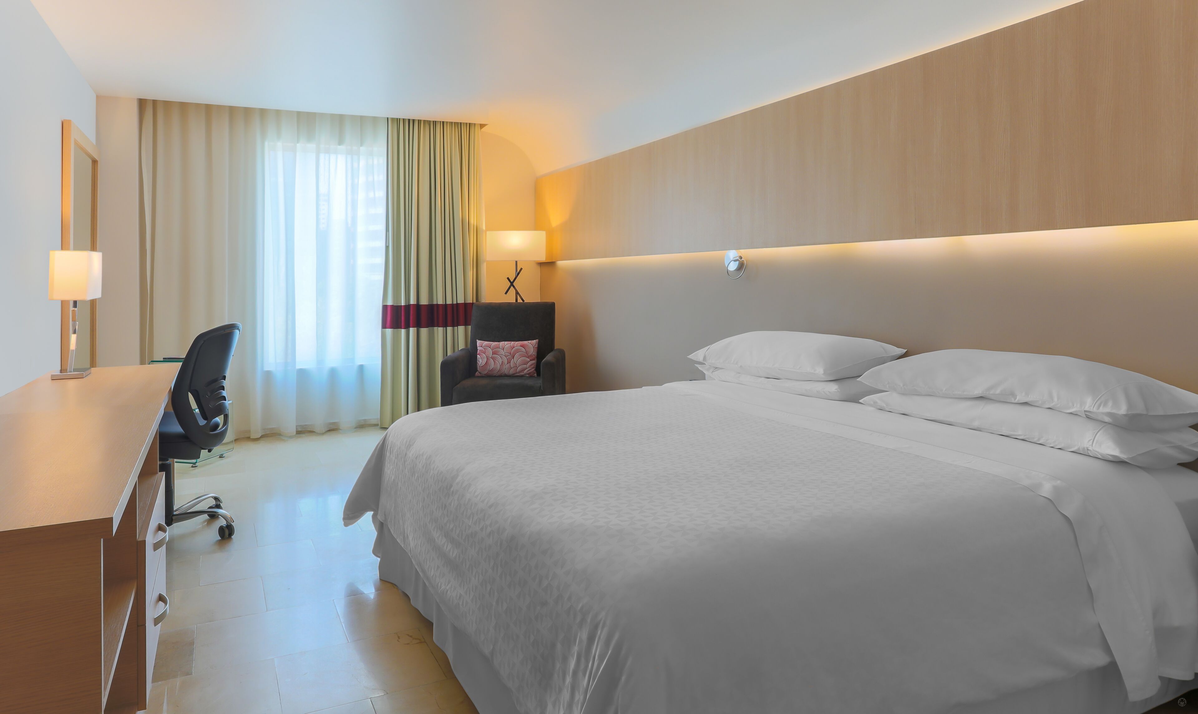 A hotel room with a large bed, a desk with a chair, a lamp, and a window with curtains.