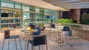 An outdoor seating area with round tables and chairs, a bar with a TV, and a glass wall overlooking the interior. - Four Points By Sheraton Barranquilla (Barranquilla)
