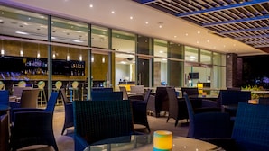 A modern outdoor seating area with glass tables and wicker chairs, a bar with a variety of bottles, and a large windowed wall providing views. - Four Points By Sheraton Barranquilla (Barranquilla)