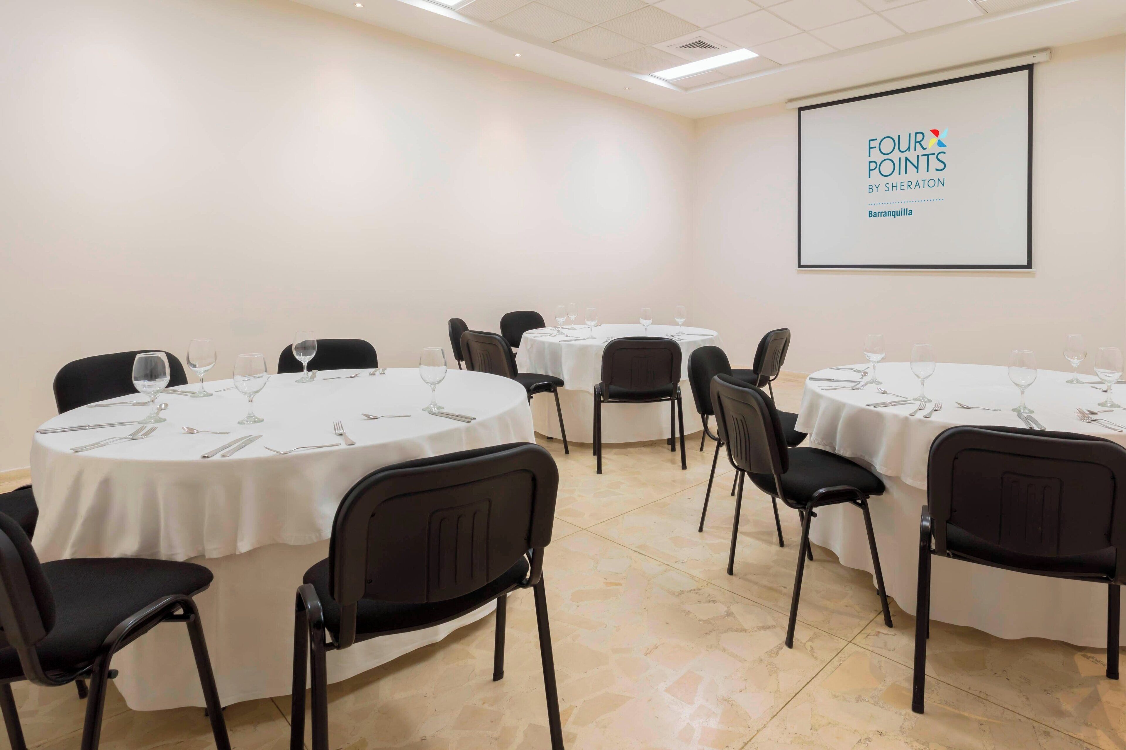 A conference room with multiple round tables, each set with chairs, wine glasses, and cutlery. A projector screen displays the text "FOUR POINTS BY SHERATON Barranquilla".