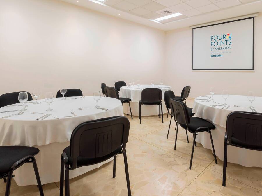 A conference room with multiple round tables, each set with chairs, wine glasses, and cutlery. A projector screen displays the text "FOUR POINTS BY SHERATON Barranquilla".