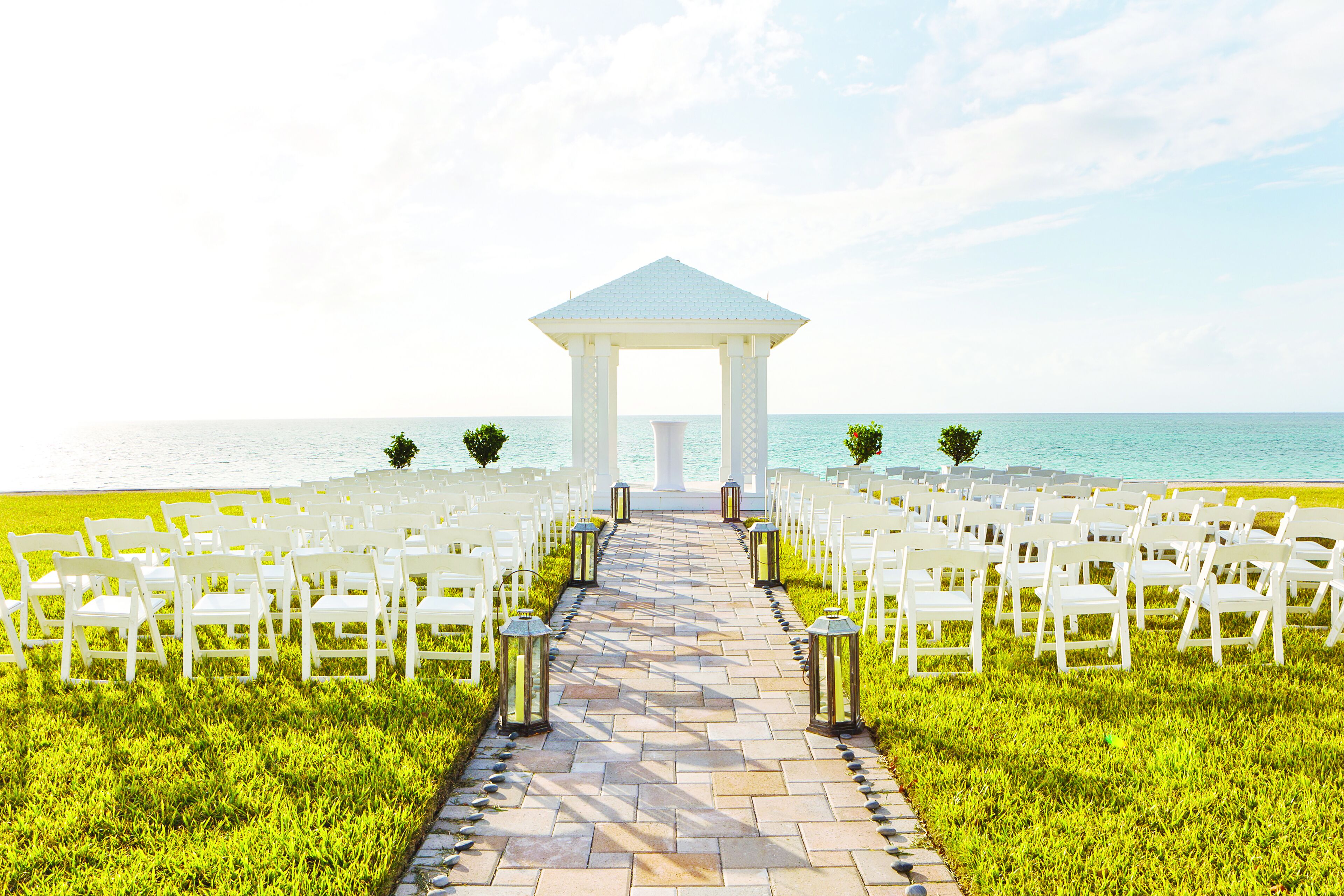 A gazebo with a tiled pathway leading to white chairs arranged on both sides, overlooking a body of water.