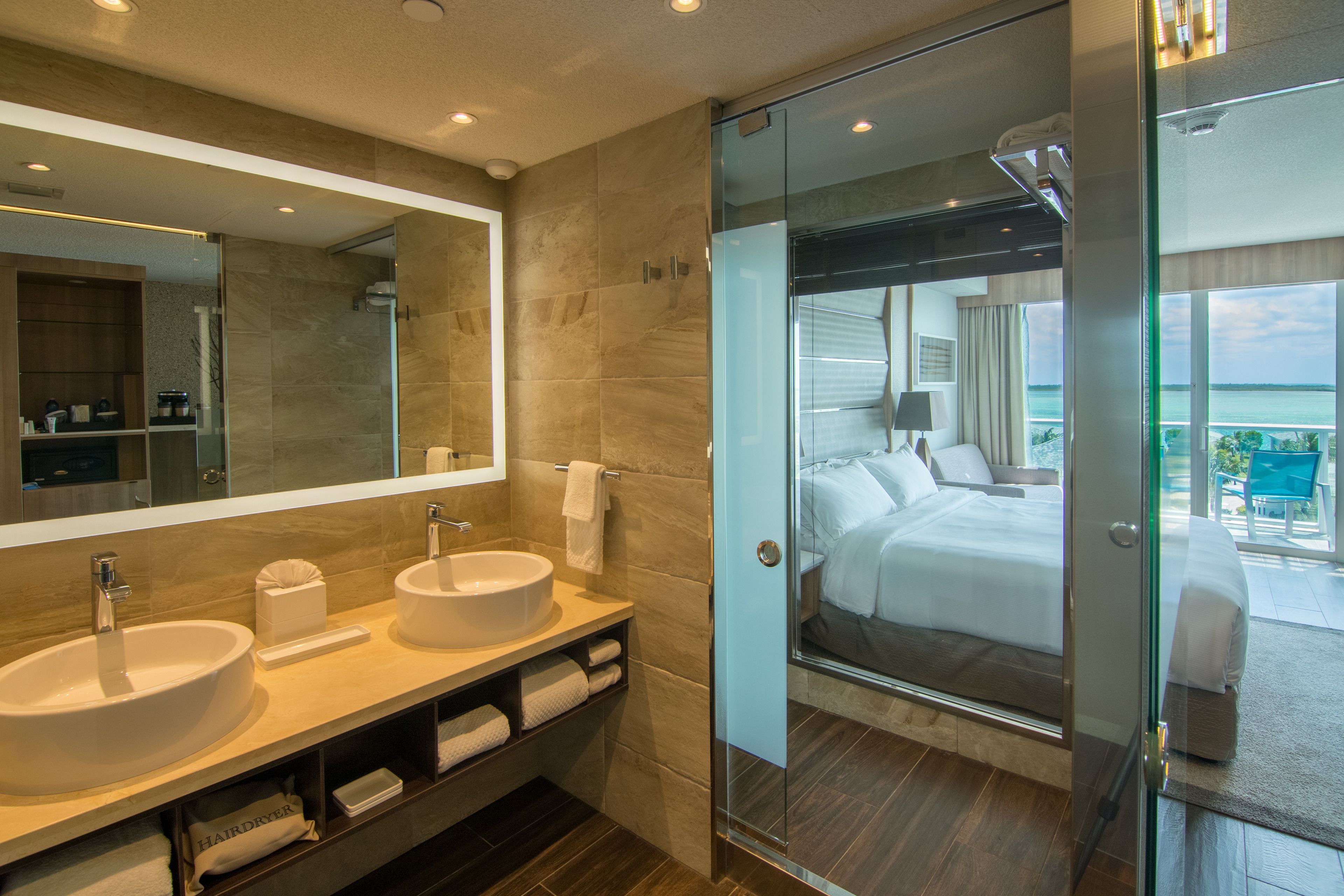 A modern bathroom with two sinks, a large mirror, and a glass-enclosed shower leading to a balcony with a bed.
