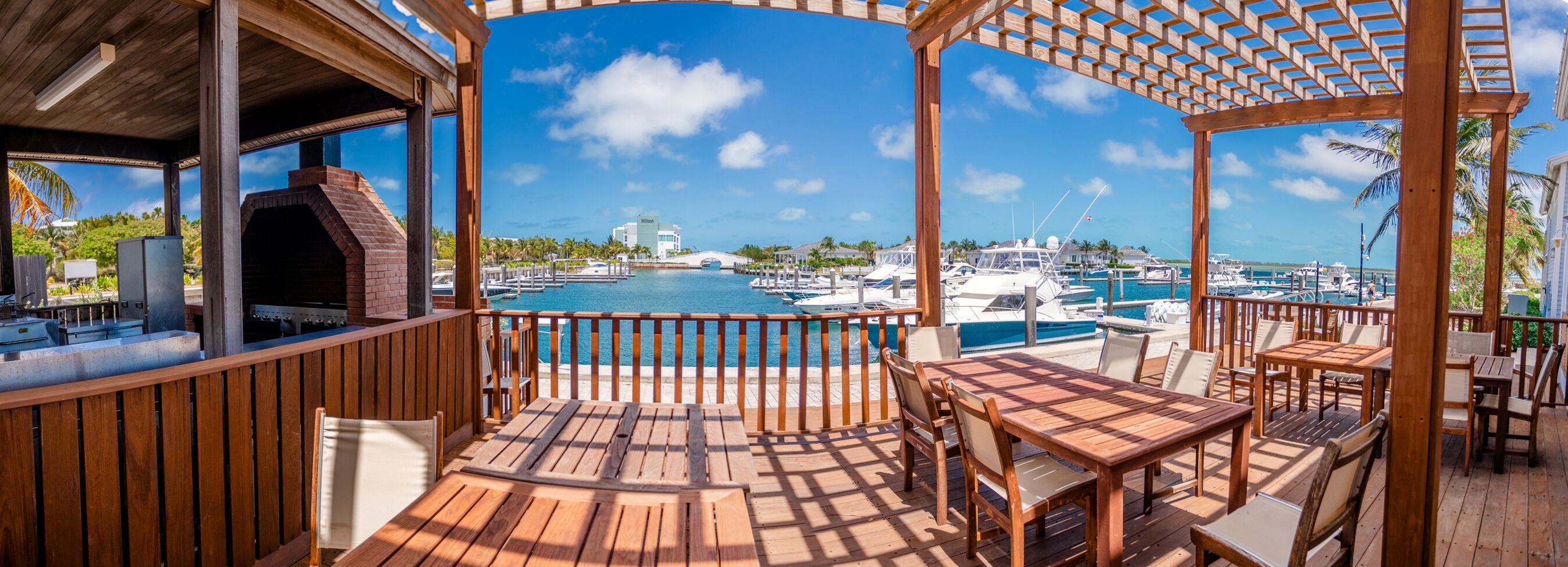 A wooden deck with outdoor seating, overlooking a marina with boats and a clear blue sky.
