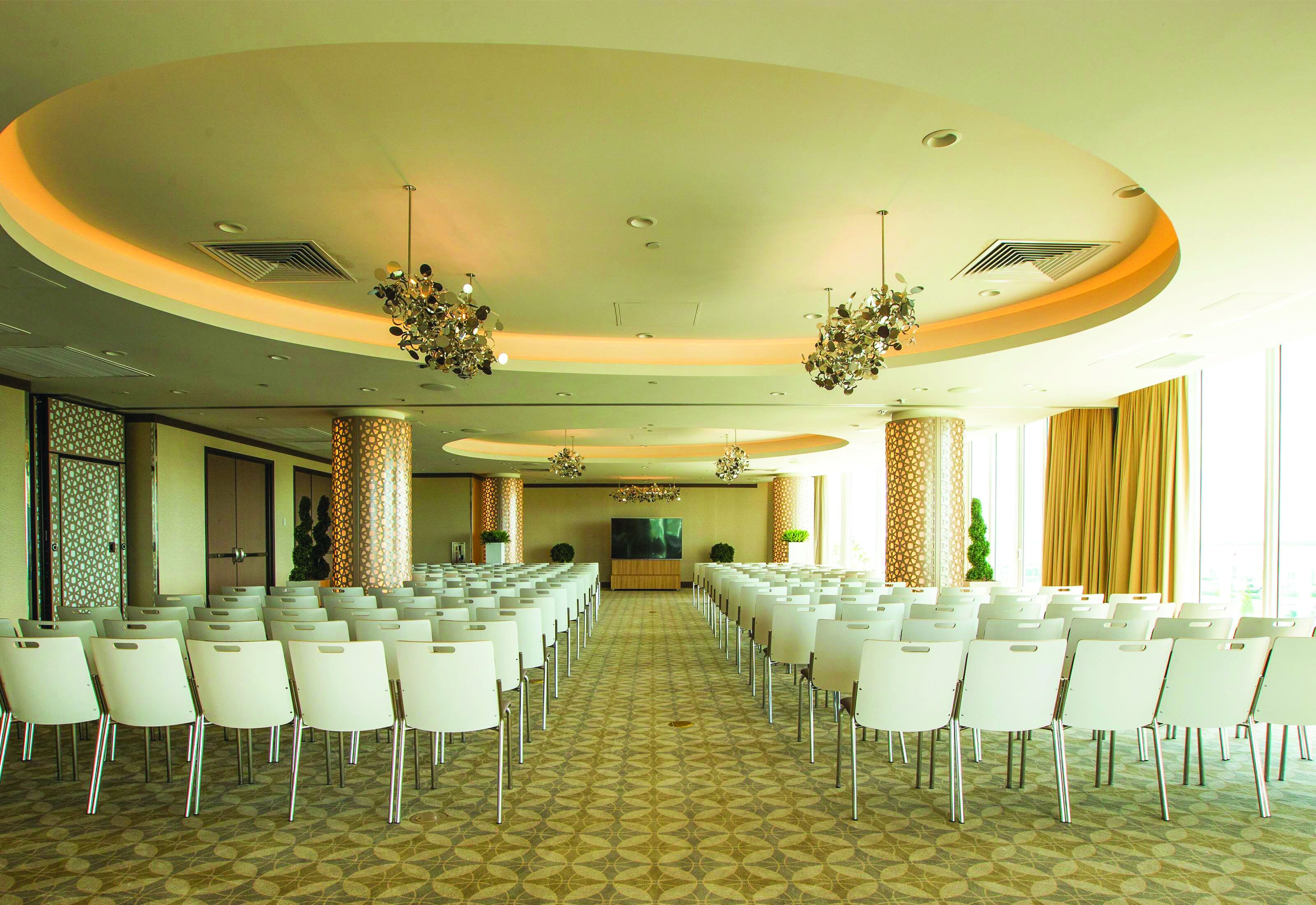 A conference room with rows of white chairs, a large screen at the front, and a patterned carpet.