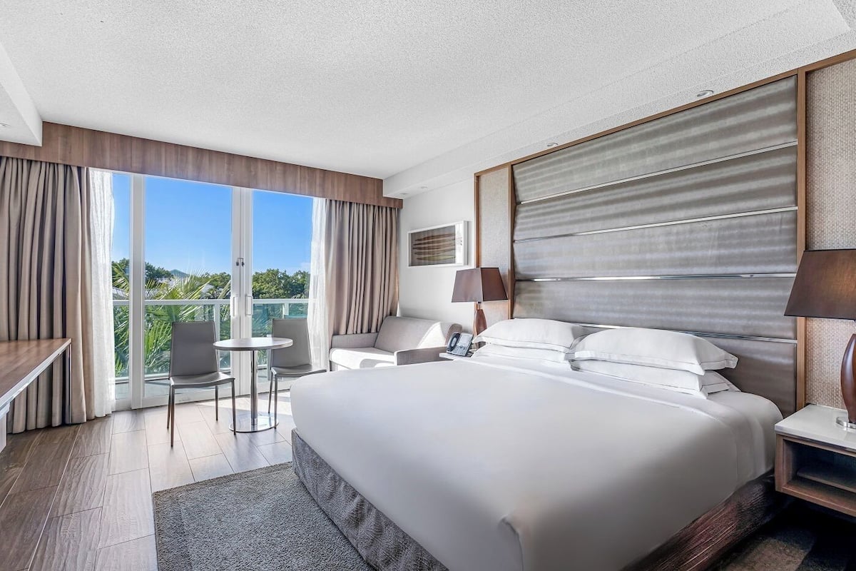 A modern hotel room with a large bed, a desk, a chair, and a balcony with a view of greenery.