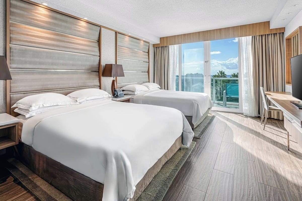 A hotel room with two beds, a desk, and a balcony with a view.