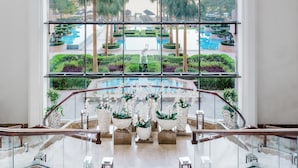Un moderno vestíbulo de hotel con una escalera de cristal, una fuente central y vistas a una zona de piscina exterior.