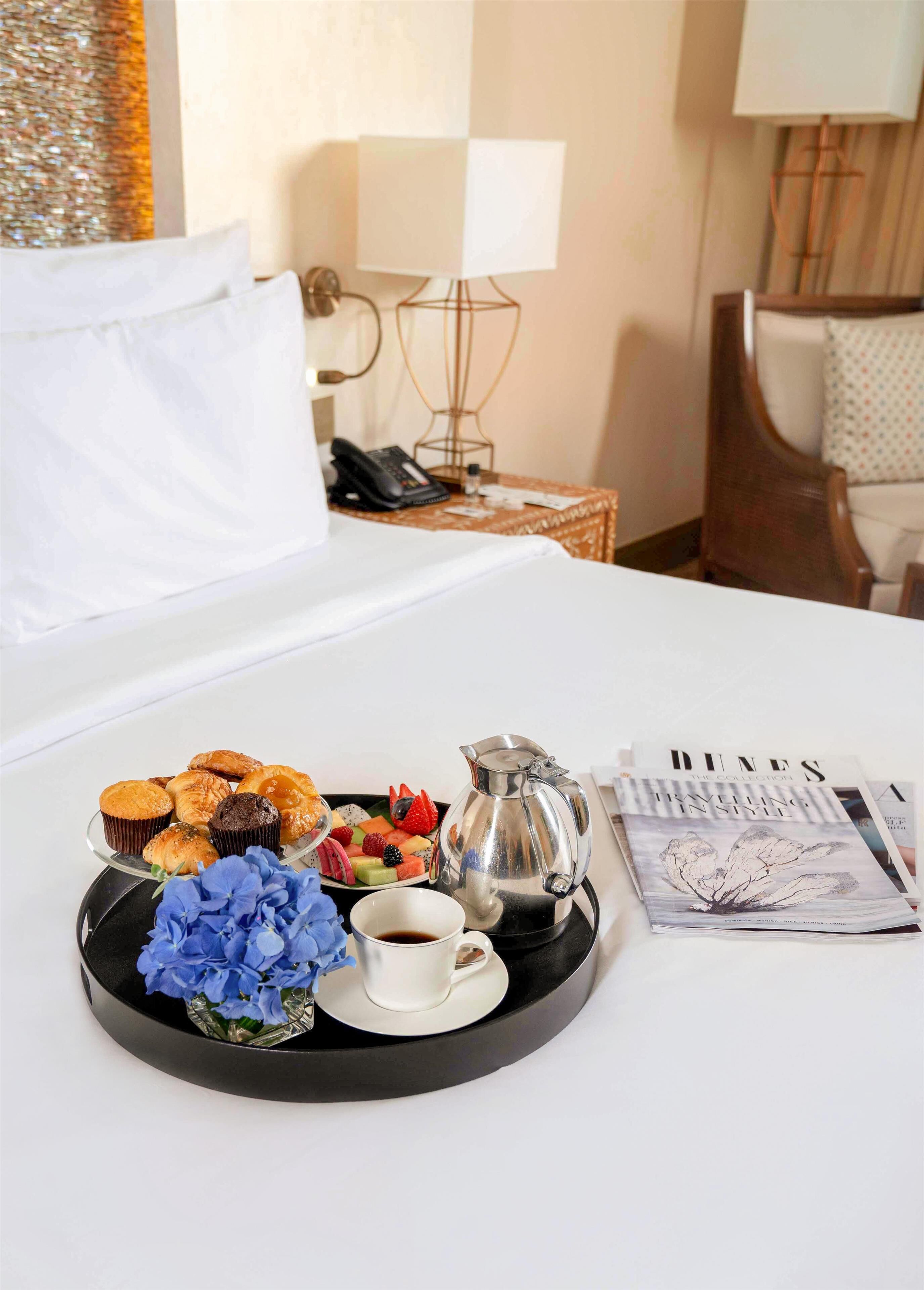Un lit bien fait, avec un plateau de viennoiseries, une cafetière et une tasse de café posés sur une table dans une chambre d’hôtel.