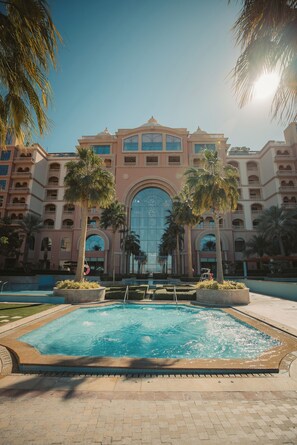 Un hotel con una gran piscina central, palmeras y una entrada imponente con ventanales abovedados.