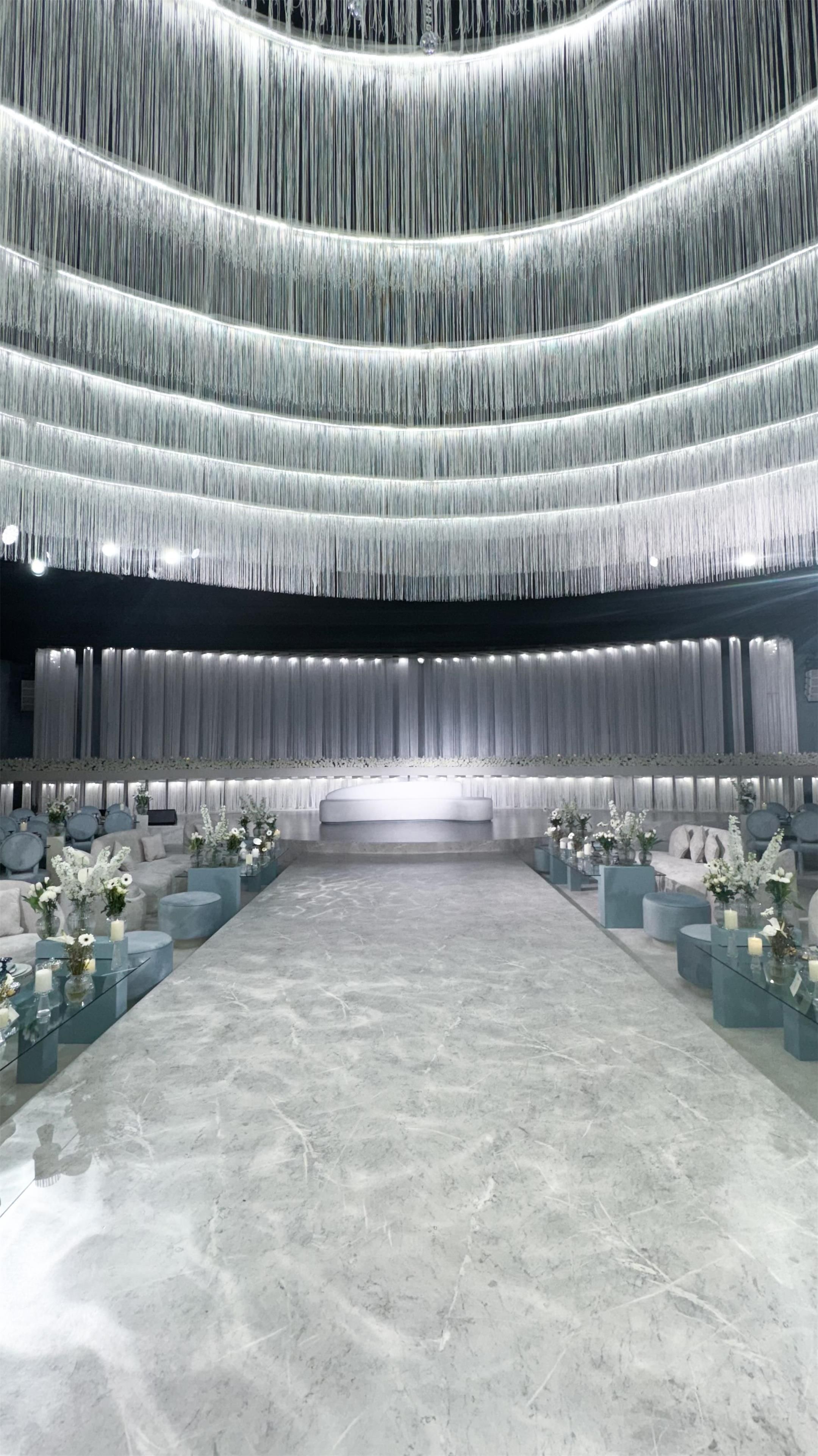 A spacious hall with tiered seating and a central stage, featuring a marble floor and floral arrangements.