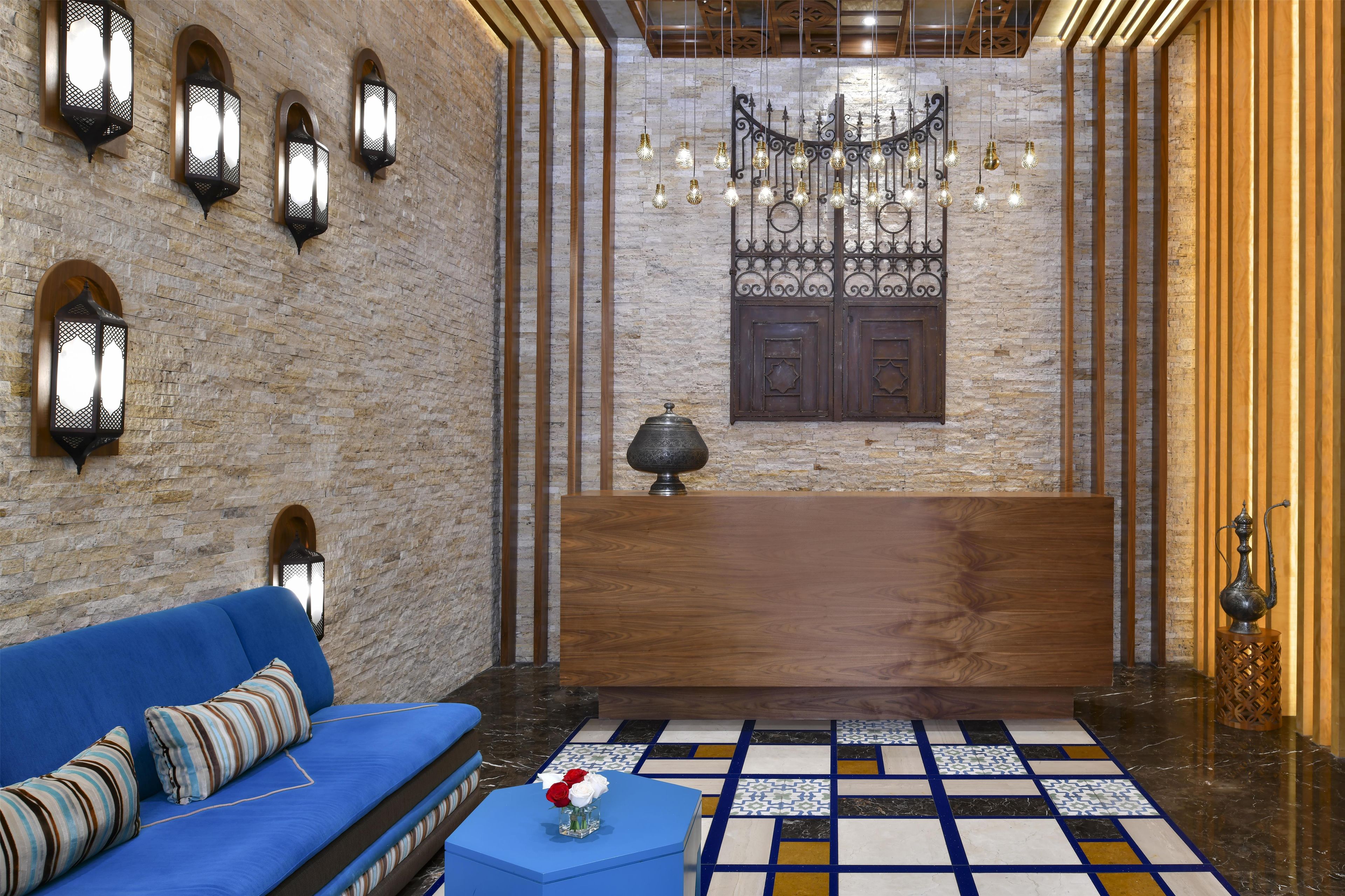 A lobby with a blue sofa, patterned cushions, a decorative lamp, a patterned floor, and a stone wall with metal fixtures.
