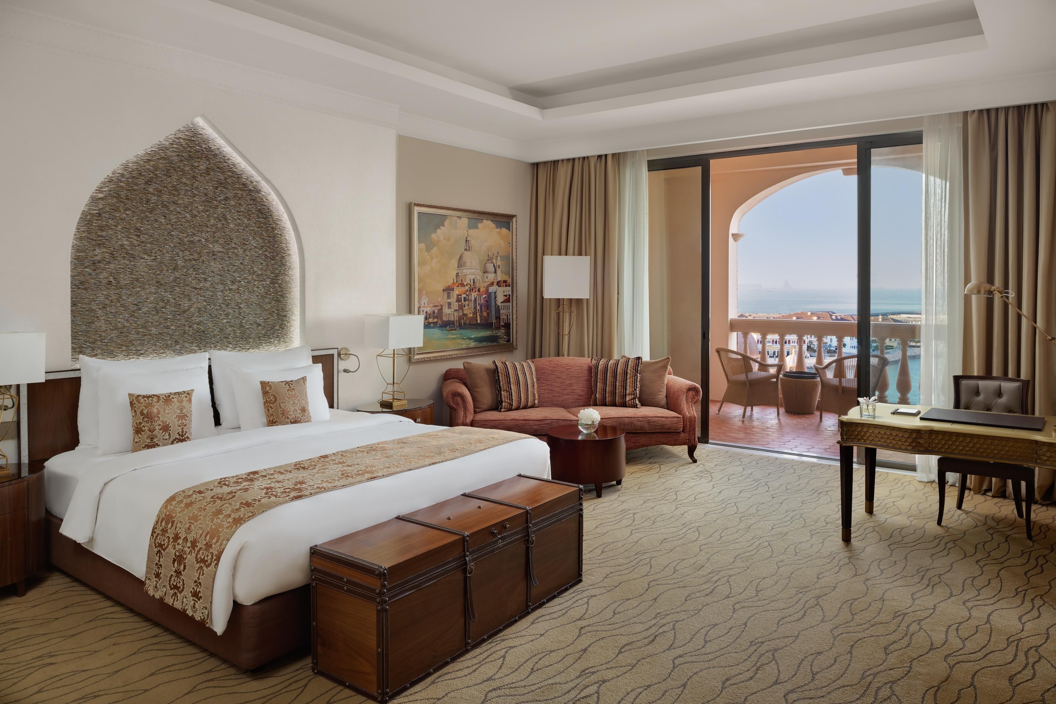 A hotel room with a large bed, a sofa, a dining table, and a view of the sea.