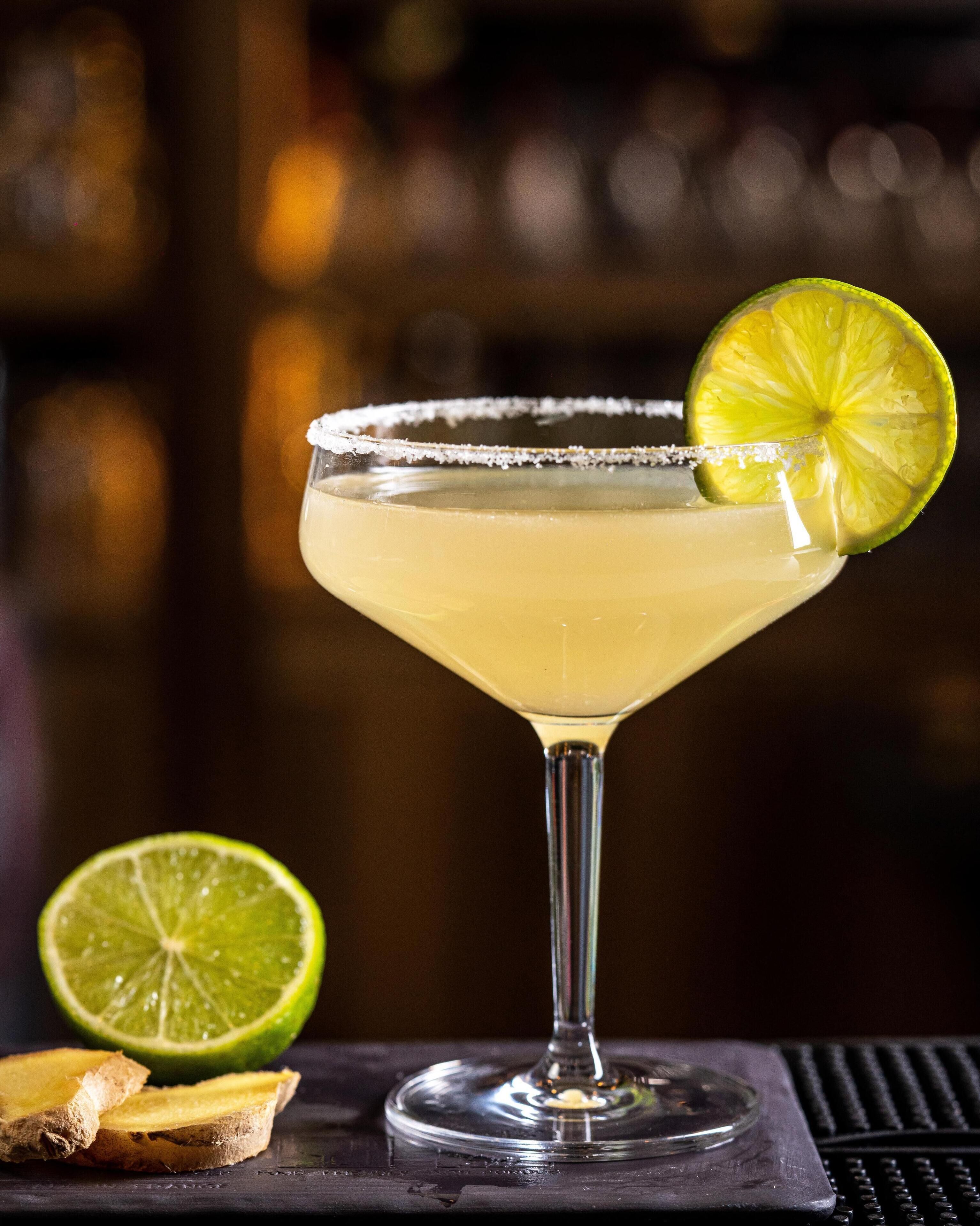 Un cocktail margarita servi dans un verre à pied, avec un bord salé et une tranche de citron vert en guise de décoration, accompagné d’un demi-citron vert et de tranches de gingembre sur une surface sombre.