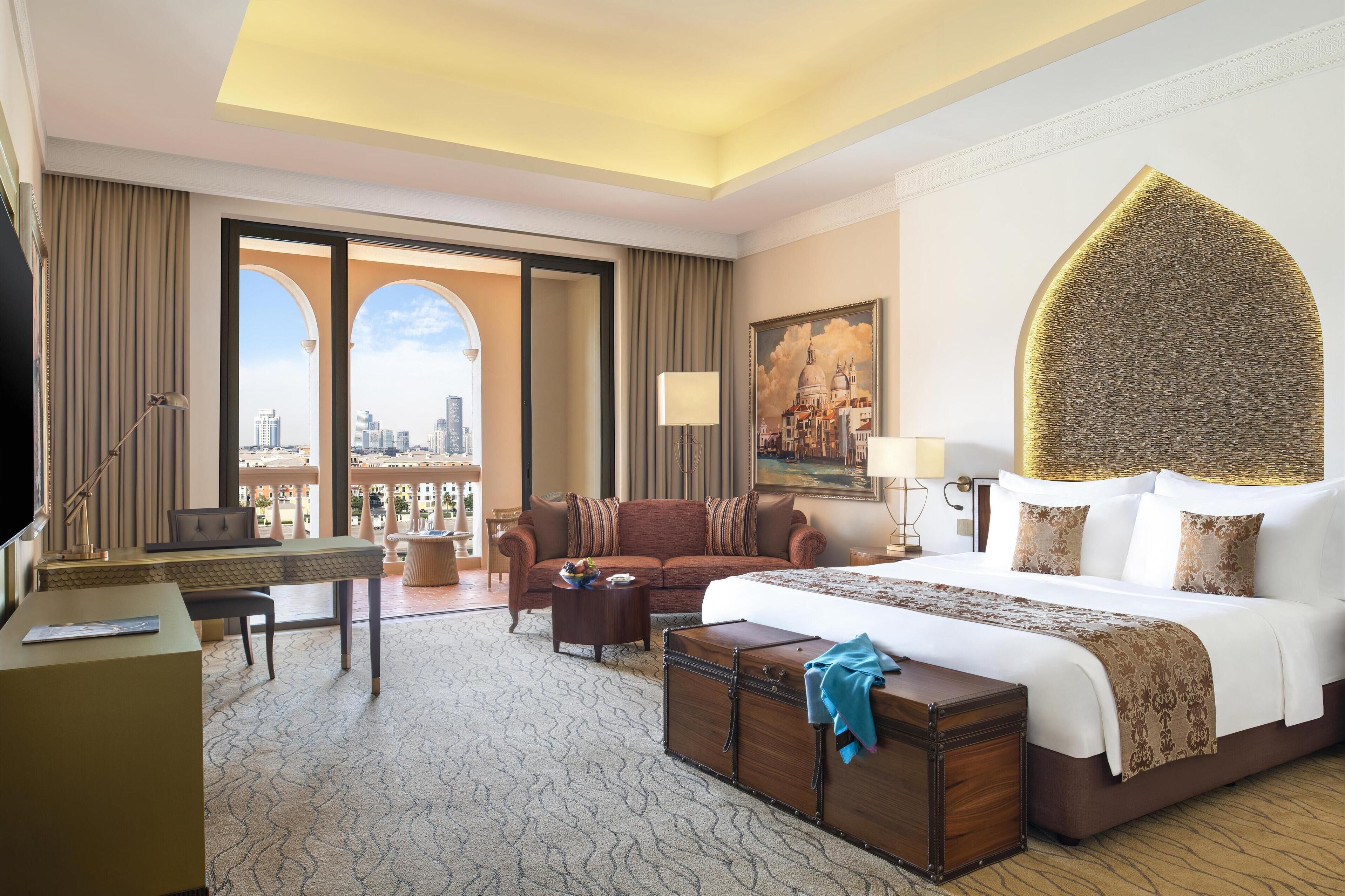 A hotel room with a large bed, a desk, a sofa, a view of the city, and a decorative wall piece.