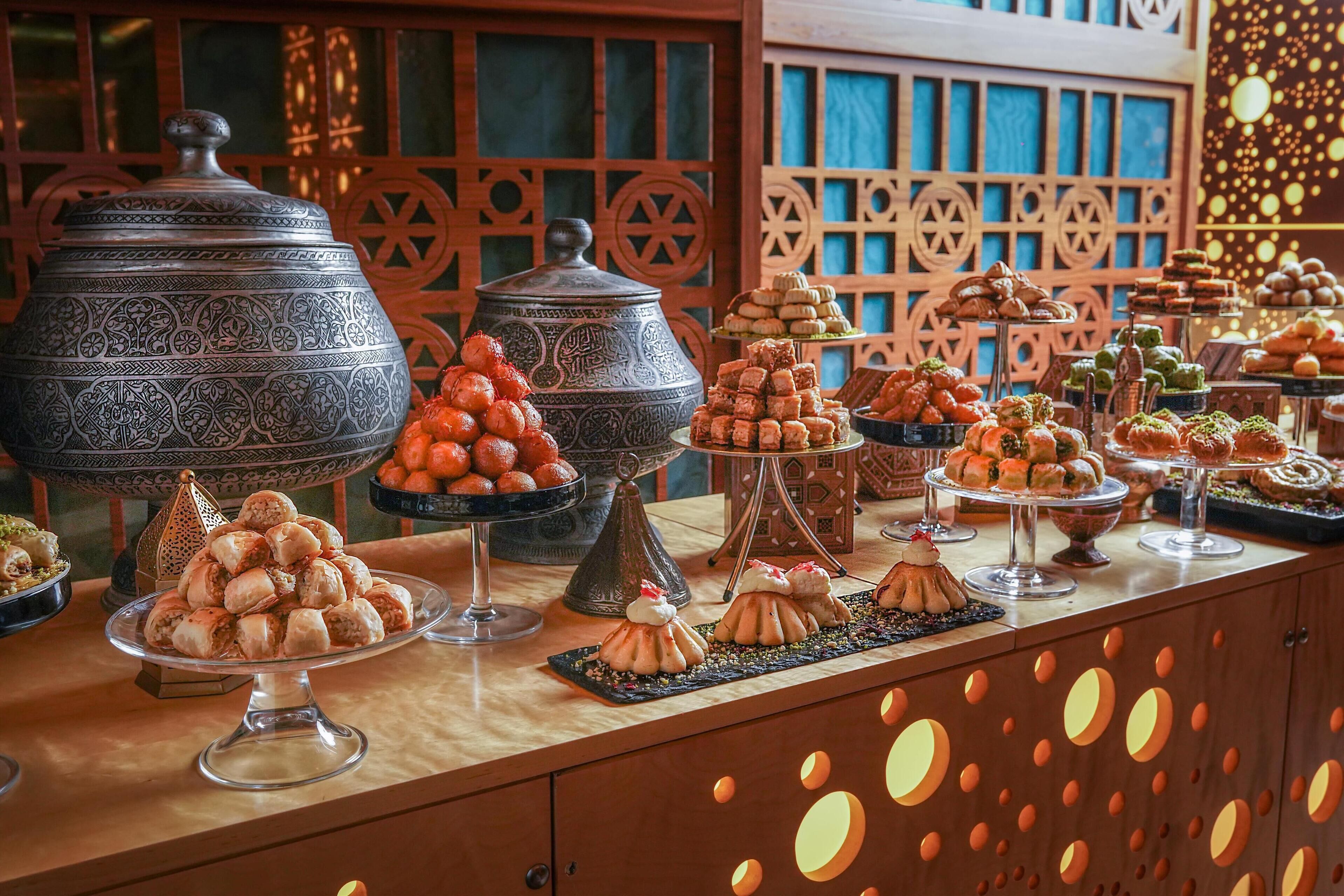 A buffet with various dishes and desserts, including a large ornate pot and a decorative wall with geometric patterns.