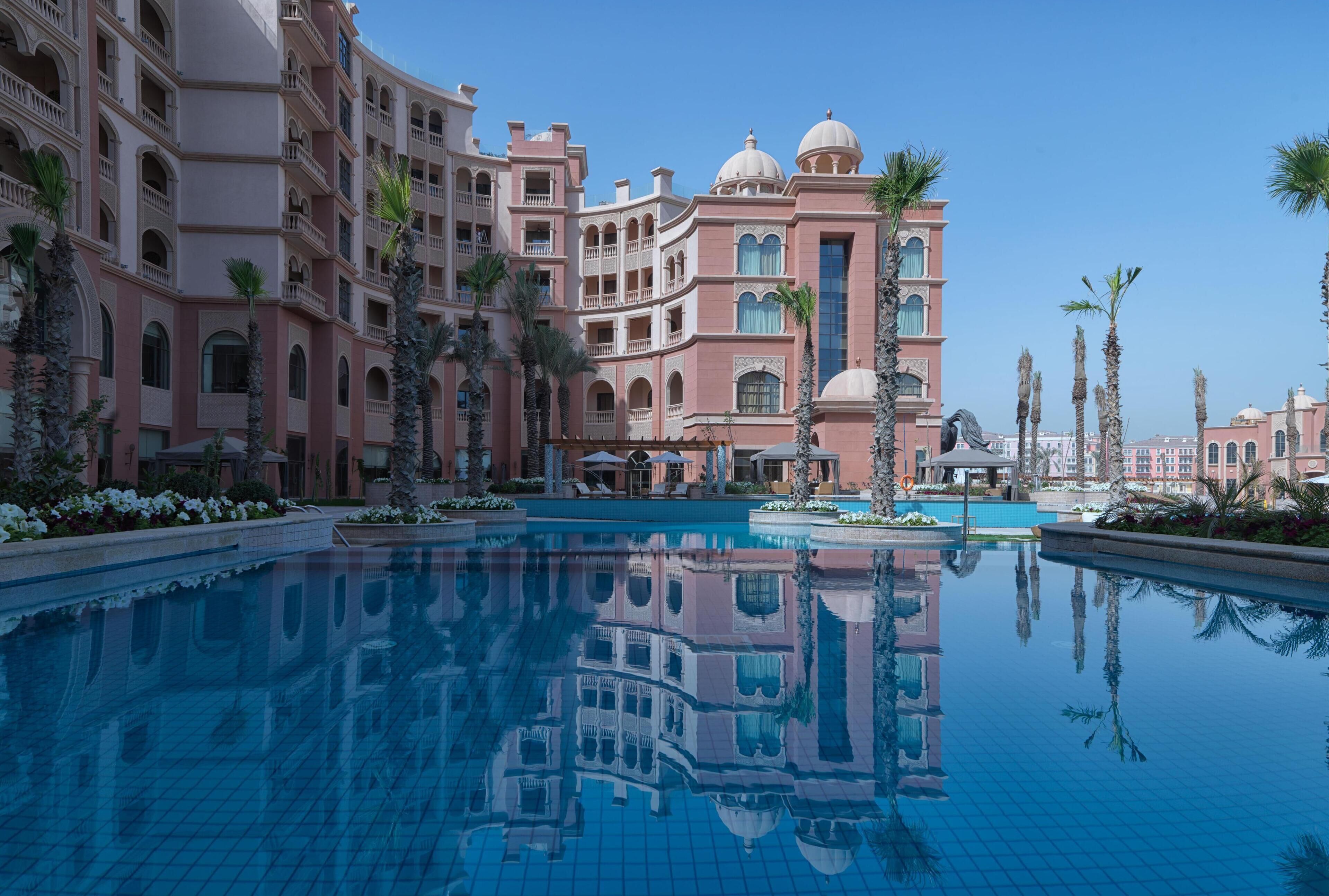 A large, clear swimming pool surrounded by a multi-story hotel with balconies and palm trees.