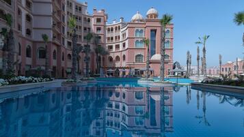 A large, clear swimming pool surrounded by a multi-story hotel with balconies and palm trees.