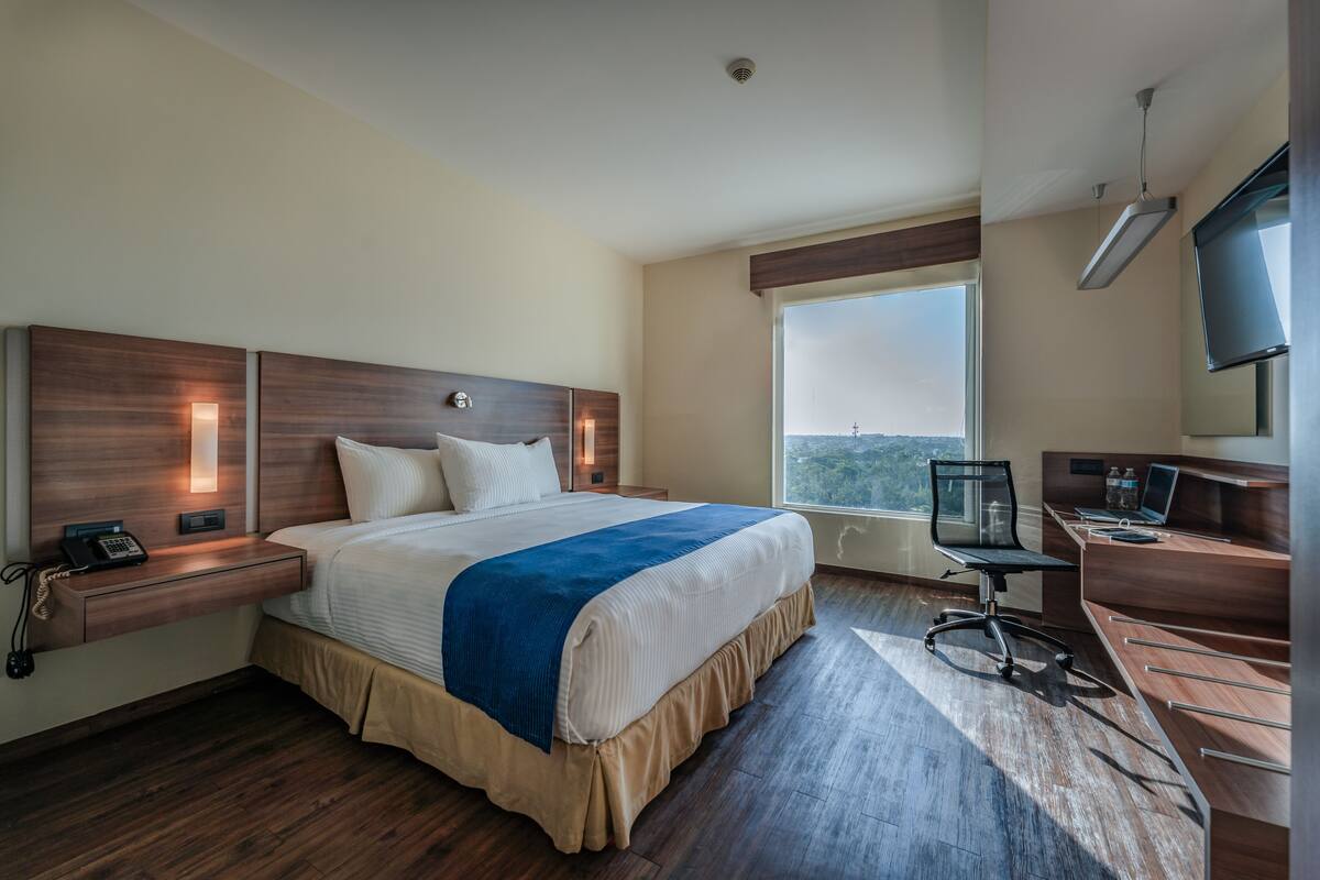 A hotel room with a large bed, a desk, a chair, a TV, and a window with a view.