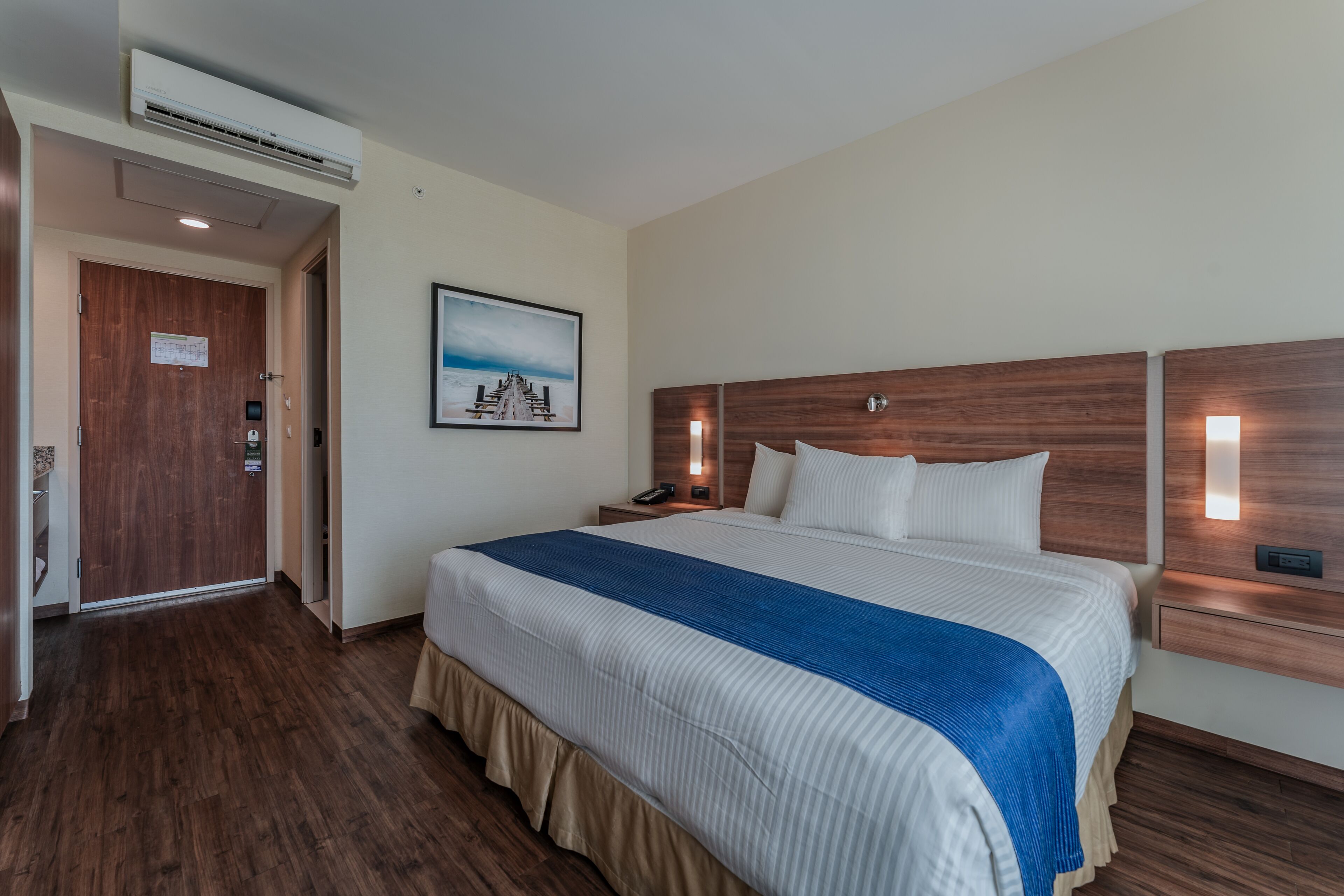 A hotel room with a large bed, wooden headboard, bedside lamps, a painting on the wall, and an air conditioning unit.