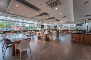 Un espacio interior amplio con varias mesas y sillas, ventanas grandes y una cocina al fondo.