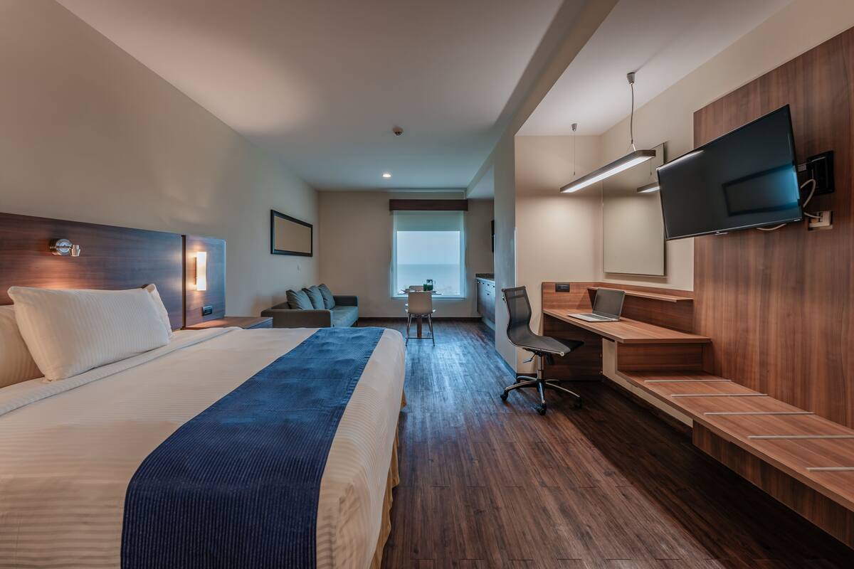 A modern hotel room with a large bed, a desk with a laptop, a TV mounted on the wall, and a view of the outdoors.