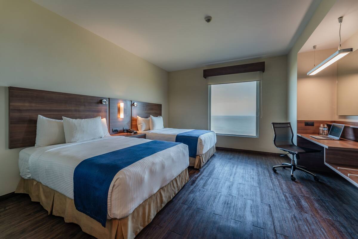 A hotel room with two beds, a desk, a chair, and a large window.