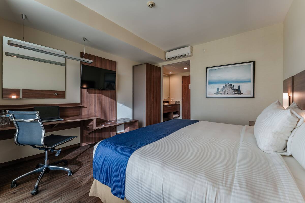 A hotel room with a bed, desk, chair, TV, and artwork on the wall.