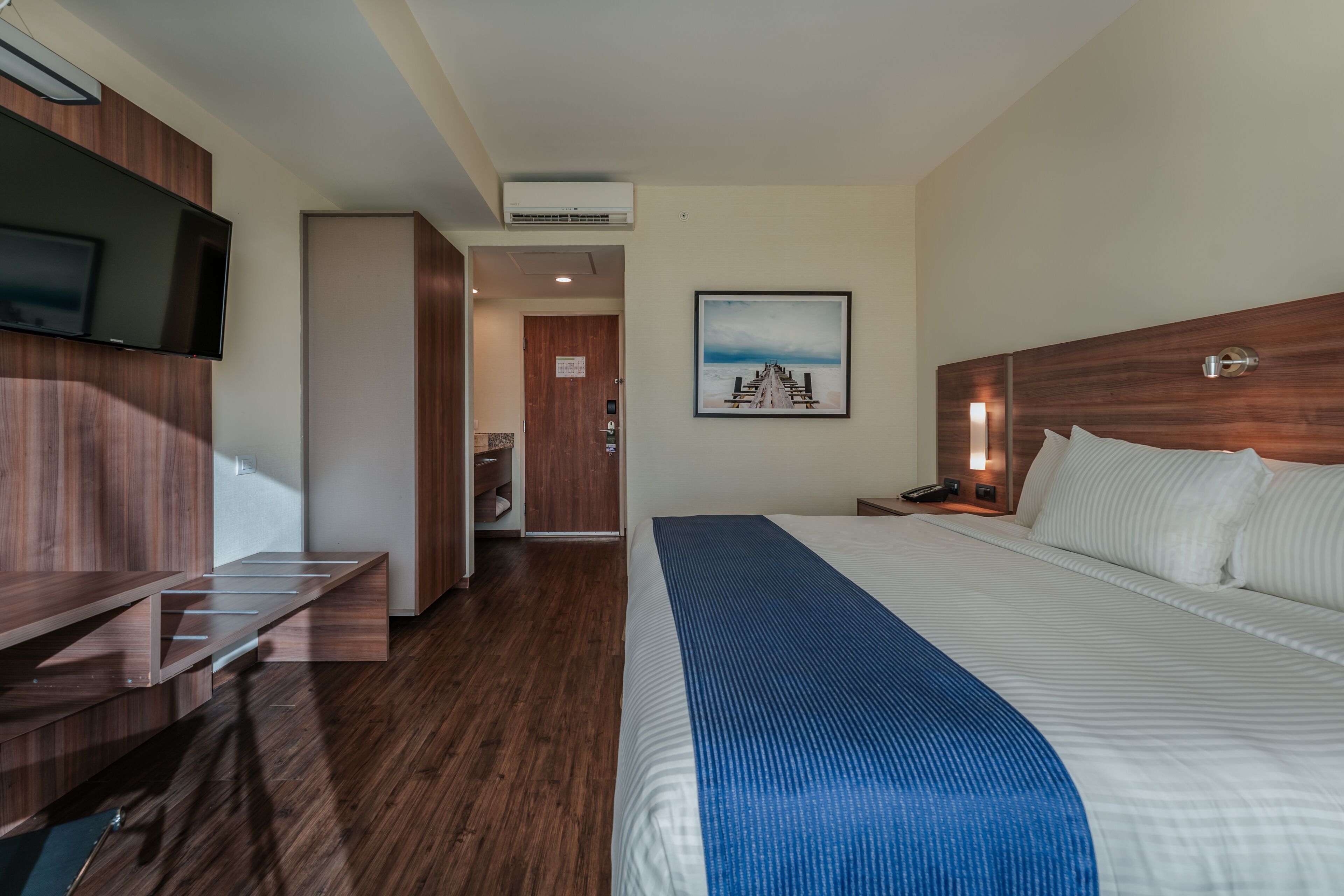 Habitación de hotel con cama, televisor montado en la pared, escritorio de madera y una imagen de un muelle.