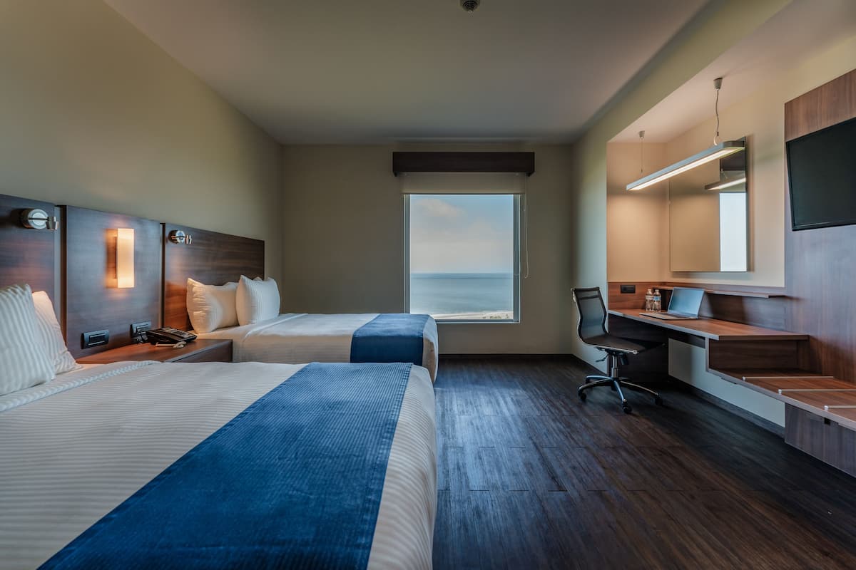 A hotel room with two beds, a desk, a chair, a large window with a view of the sea, and a mounted TV.
