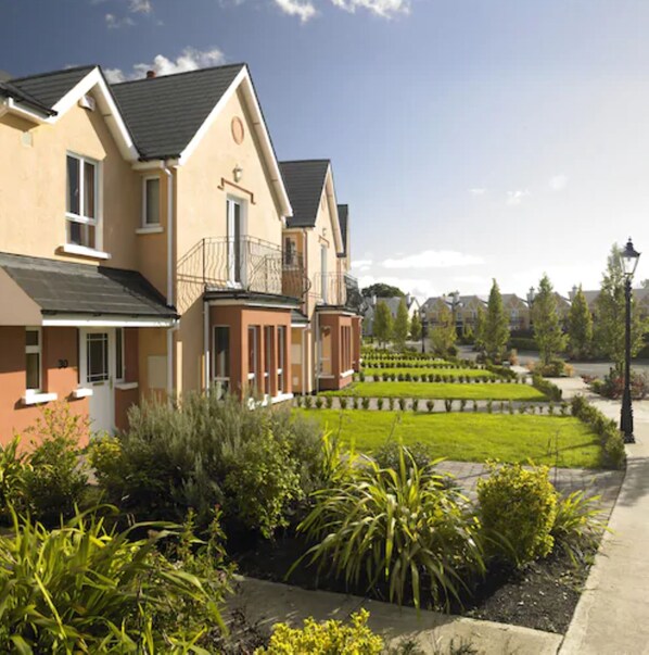 A two-story residential building with a balcony, a well-maintained garden, and a clear sky.