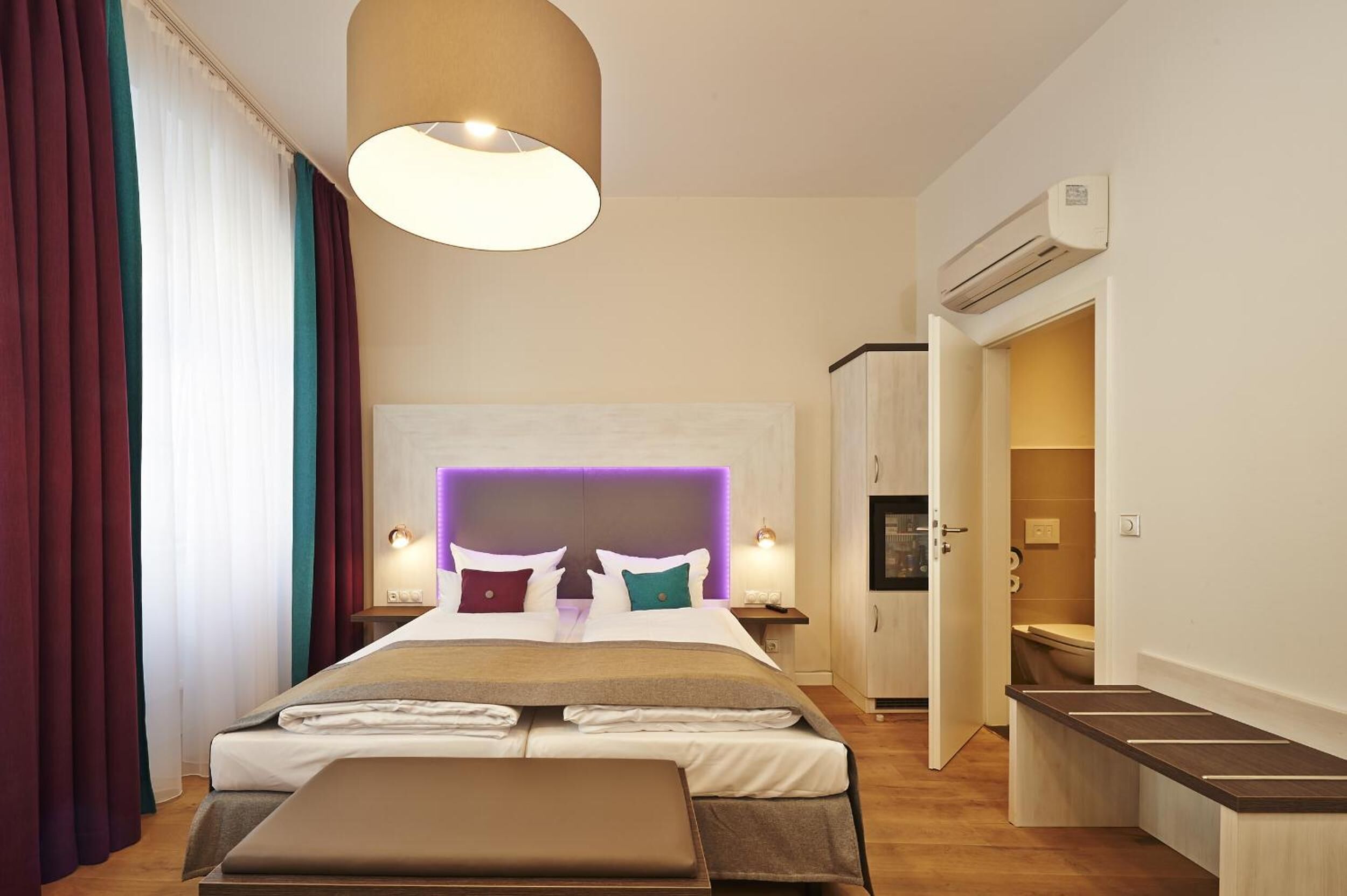 Een moderne hotelkamer met een groot bed, nachtlampjes, een hoofdeinde met ingebouwde verlichting, een airconditioning en een zichtbare badkamer door een geopende deur.